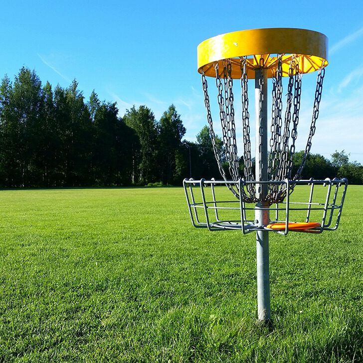 &bdquo;Disc-Golf-Korb mit gelbem oberen Ring und Metallketten steht auf einer gr&uuml;nen Wiese unter blauem Himmel. Im Hintergrund sind B&auml;ume am Rand des Spielfelds zu sehen.&ldquo;