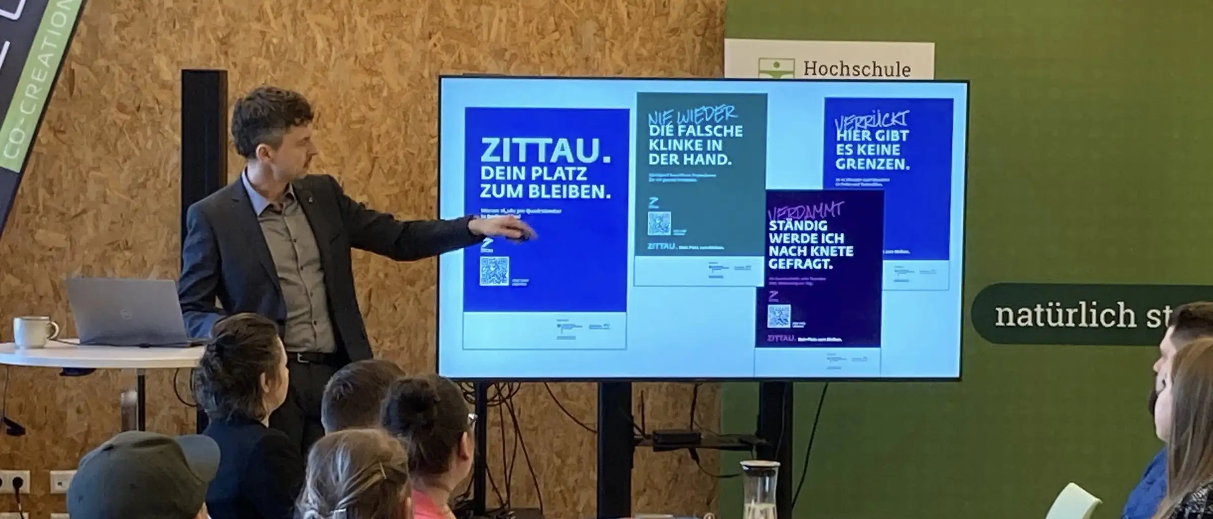 Redner Kai Grebasch zeigt auf die Kampagnenmotive auf dem Bildschirm. Im Vordergrund sitzen die Unternehmerinnen und Unternehmer und h&ouml;ren dem Vortrag zu