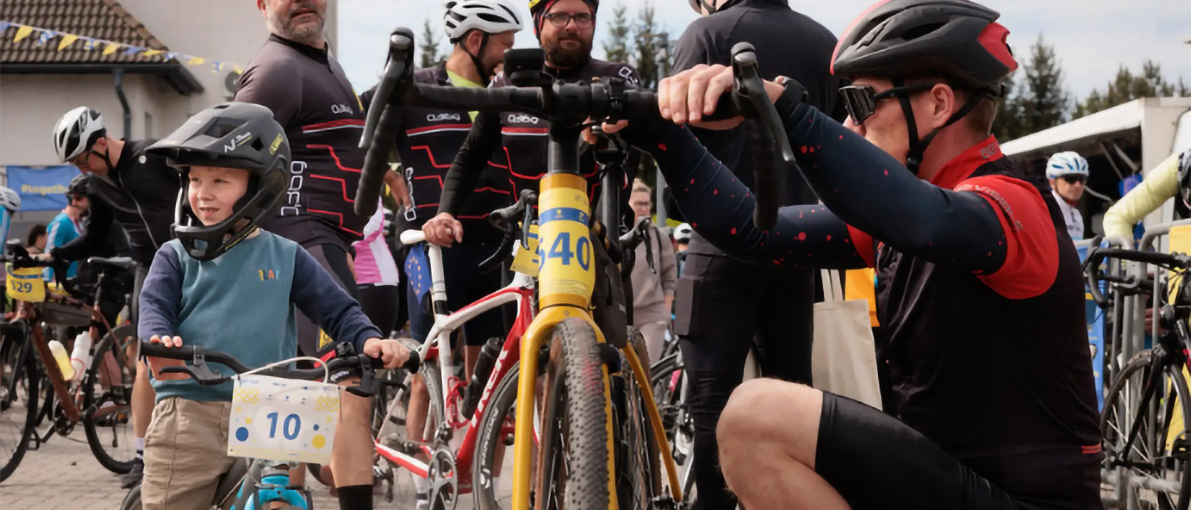 Ein Mann in Radbekleidung und Helm kniet neben seinem Fahrrad mit der Startnummer 540 und &uuml;berpr&uuml;ft den Lenker. Neben ihm steht ein kleiner Junge mit Helm und der Startnummer 10 auf seinem Kinderfahrrad. Im Hintergrund bereiten sich weitere Radfahrer auf die Tour vor, einige tragen Trikots mit roten Streifen. Die Szene spielt auf einem Platz vor einem Geb&auml;ude bei Tageslicht und vermittelt Vorfreude auf die gemeinsame Radtour.