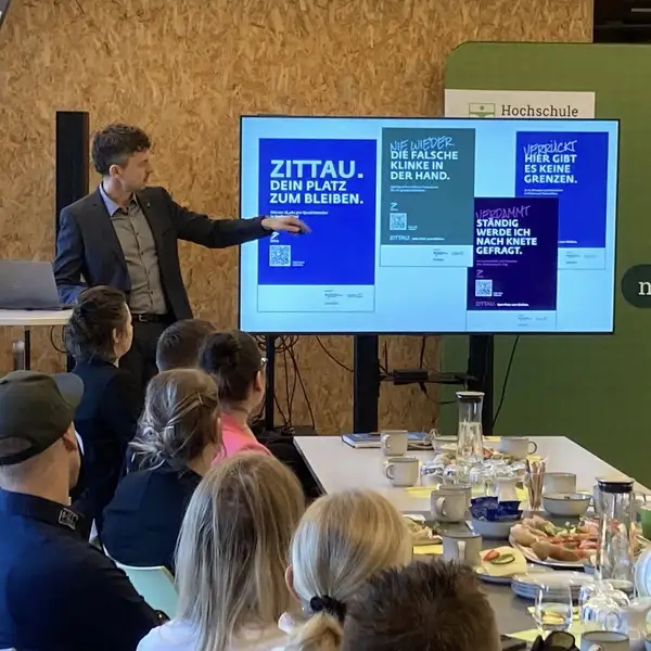 Redner Kai Grebasch zeigt auf die Kampagnenmotive auf dem Bildschirm. Im Vordergrund sitzen die Unternehmerinnen und Unternehmer und h&ouml;ren dem Vortrag zu