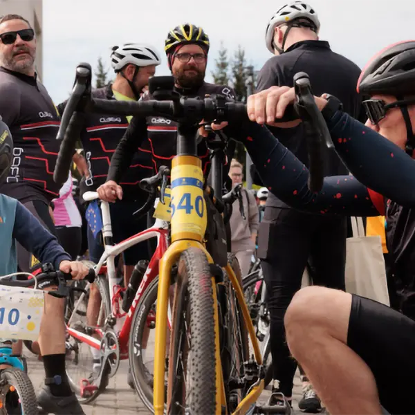 Ein Mann in Radbekleidung und Helm kniet neben seinem Fahrrad mit der Startnummer 540 und &uuml;berpr&uuml;ft den Lenker. Neben ihm steht ein kleiner Junge mit Helm und der Startnummer 10 auf seinem Kinderfahrrad. Im Hintergrund bereiten sich weitere Radfahrer auf die Tour vor, einige tragen Trikots mit roten Streifen. Die Szene spielt auf einem Platz vor einem Geb&auml;ude bei Tageslicht und vermittelt Vorfreude auf die gemeinsame Radtour.